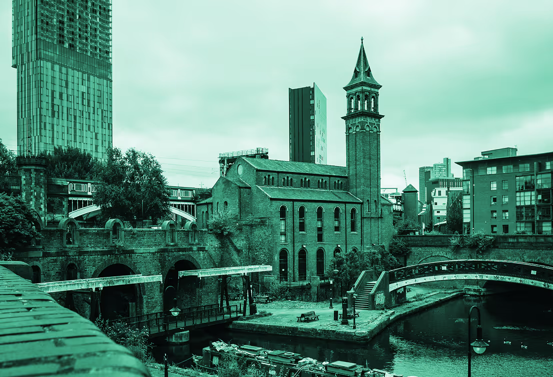 Urban canal scene with historic brick buildings, industrial bridges, and modern skyscrapers in the background under a cloudy sky.